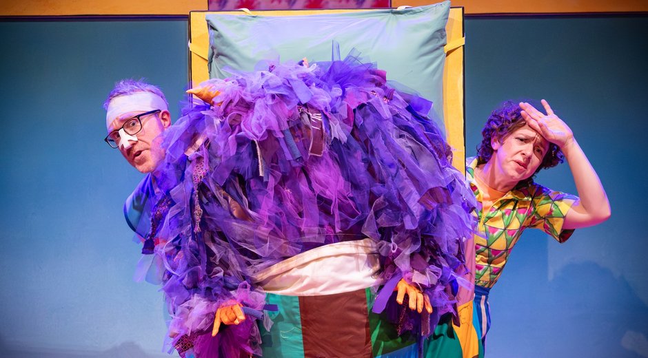 Two people peeping out behind a purple fluffy monster